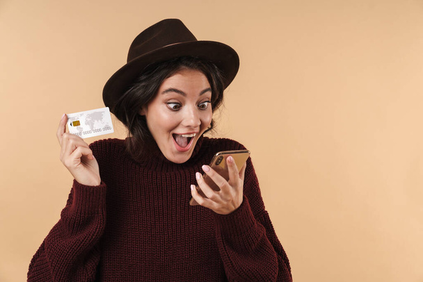 Image of screaming happy positive young brunette woman posing isolated over beige wall background holding credit card using mobile phone. - Fotografie, Obrázek