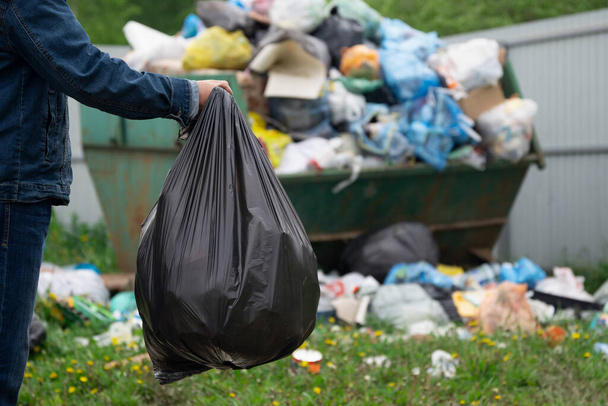 Un homme en costume de denim jette un sac en plastique avec une poubelle à la poubelle à l'extérieur à la campagne. Concept d'élimination des ordures. - Photo, image