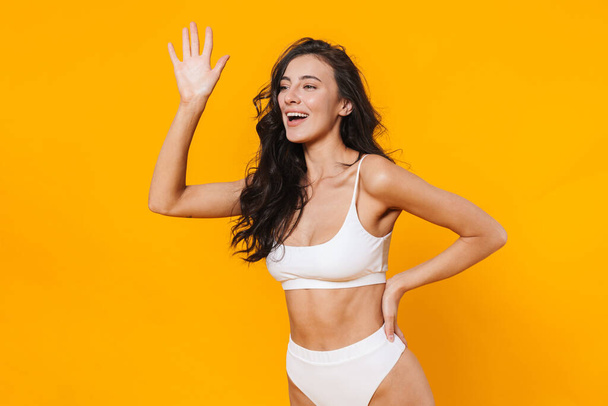 Image of beautiful joyful woman in swimsuit smiling and waving hand isolated over yellow wall - Foto, Bild