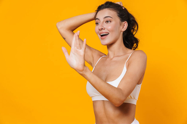 Image of beautiful joyful woman in swimsuit smiling and waving hand isolated over yellow wall - Photo, Image