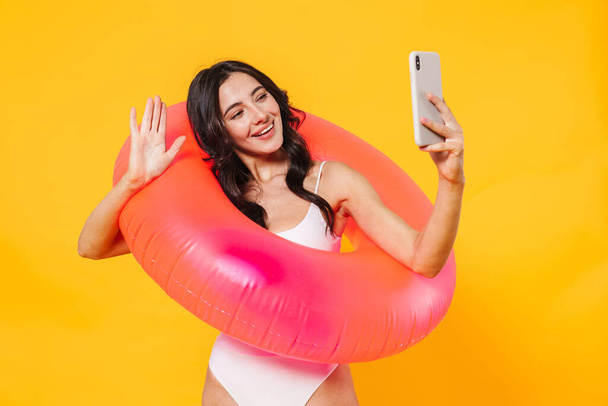 Image of cheerful woman with rubber ring waving hand and taking selfie on cellphone isolated over yellow wall - Foto, afbeelding