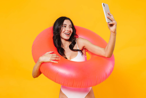 Image of cheerful woman with rubber ring smiling and taking selfie on cellphone isolated over yellow wall - Photo, Image