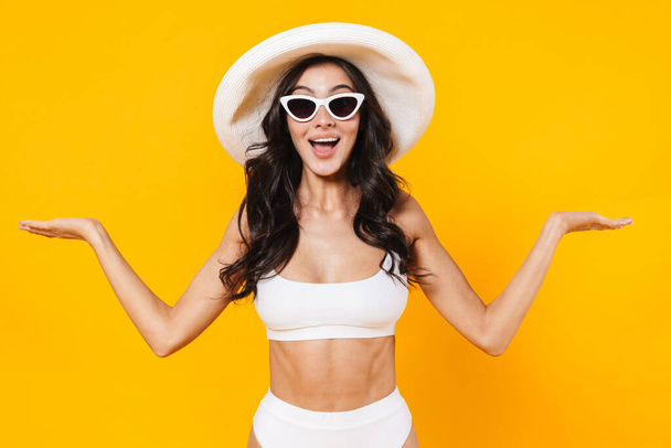 Image of stylish delighted woman in swimsuit and hat smiling and holding copyspace isolated over yellow wall - Foto, immagini