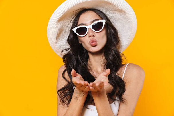 Image of stylish young woman in swimsuit and hat blowing air kiss at camera isolated over yellow wall - Foto, Imagem