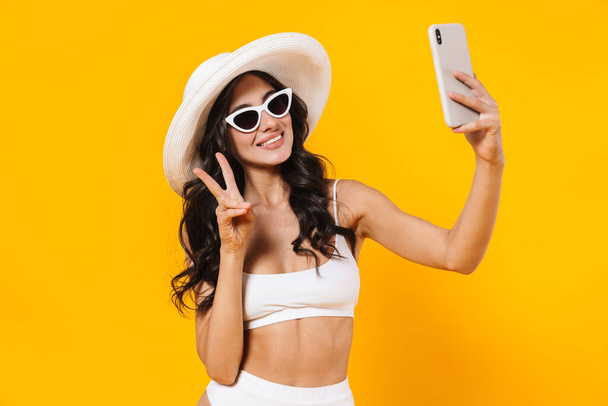 Image of cheerful woman in swimsuit and hat gesturing peace sign while taking selfie on cellphone isolated over yellow wall - Fotó, kép