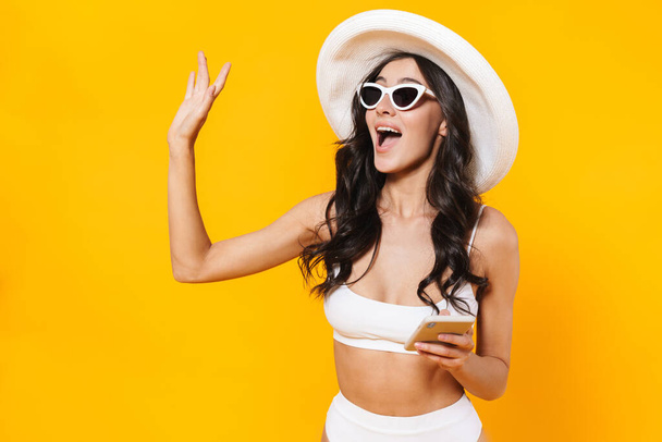 Image of elegant cheerful woman in swimsuit and hat waving hand and using cellphone isolated over yellow wall - Foto, Imagem