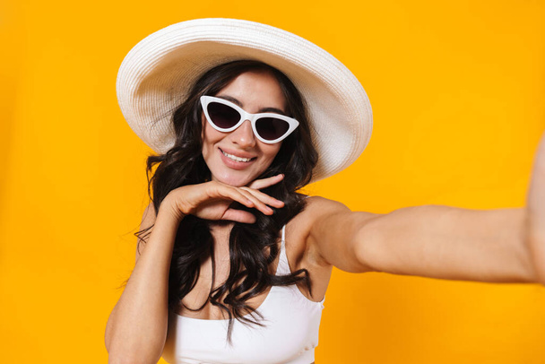 Image of cheerful woman in swimsuit and hat smiling while taking selfie photo isolated over yellow wall - Fotoğraf, Görsel