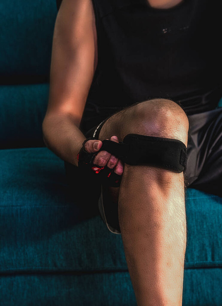 Vista frontal de un hombre cerrando una correa de rodilla sentado en un sofá antes de entrenar en casa
 - Foto, Imagen