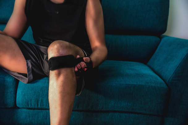 Front view of a man closing a knee strap sitting on a couch before home workout - Photo, Image