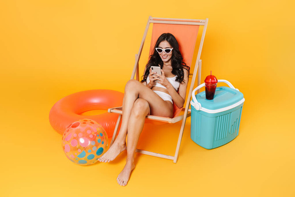 Image of happy woman in swimsuit using smartphone while resting in chaise lounge isolated over yellow wall - Фото, изображение