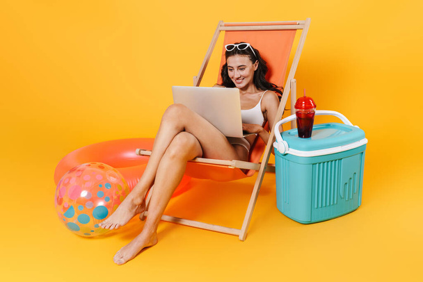 Image of happy woman in swimsuit using laptop while resting in chaise lounge isolated over yellow wall - Zdjęcie, obraz