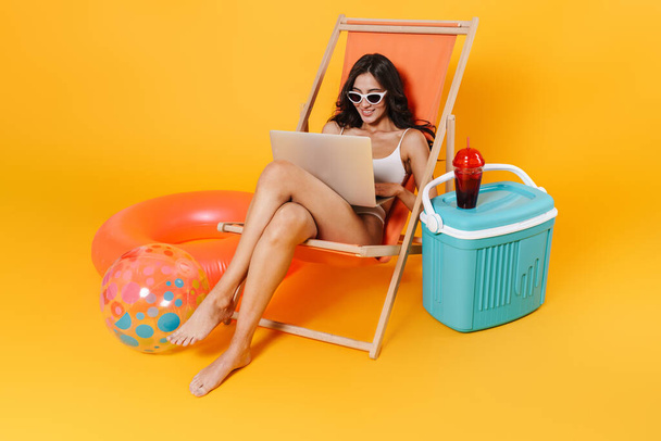 Image of happy woman in swimsuit using laptop while resting in chaise lounge isolated over yellow wall - Фото, изображение