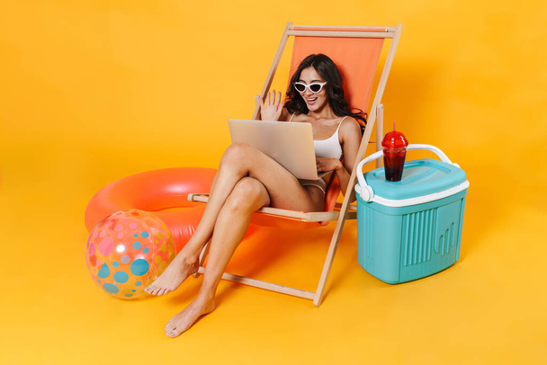 Image of happy woman in swimsuit using laptop and waving hand while resting in chaise lounge isolated over yellow wall - Foto, immagini