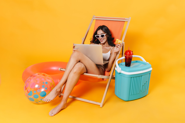 Image of happy woman using laptop and holding credit card while resting in chaise lounge isolated over yellow wall - Fotografie, Obrázek
