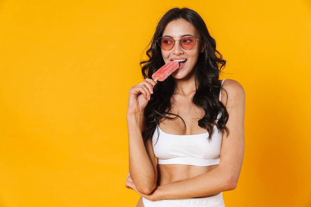 Image of attractive nice woman in swimsuit and eyeglasses eating ice cream isolated over yellow wall - Foto, Imagem
