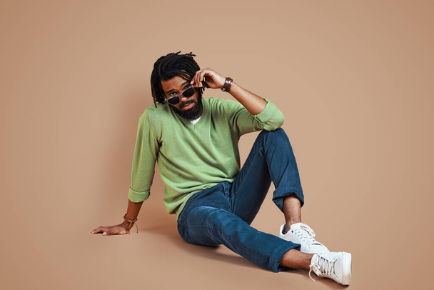 Trendy young African man in casual clothing looking at camera and adjusting eyewear while sitting against brown background - Foto, Imagem