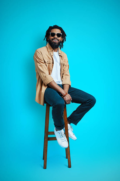 Full length of handsome young African man in casual wear looking at camera and smiling while sitting on stool against blue background                      - Zdjęcie, obraz