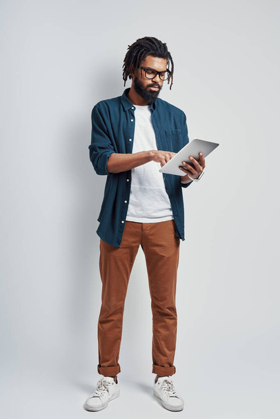 Full length of young African man in eyewear using digital tablet while standing against grey background - Valokuva, kuva