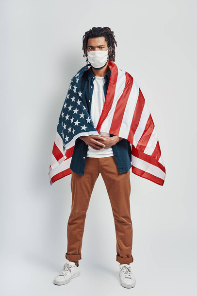 Longitud completa de hombre africano joven guapo cubierto con bandera americana con máscara facial médica y mirando a la cámara mientras está de pie sobre fondo gris
          - Foto, Imagen