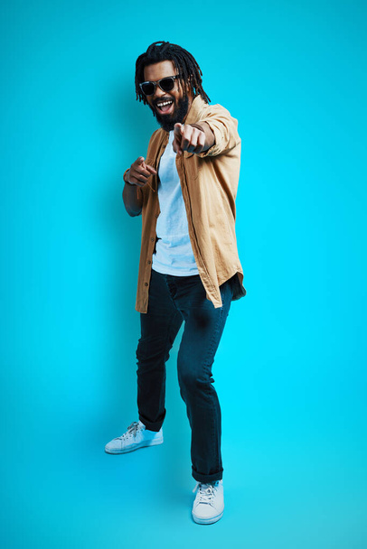 Full length of handsome young African man in casual wear looking at camera and pointing you while standing against blue background              - Fotoğraf, Görsel