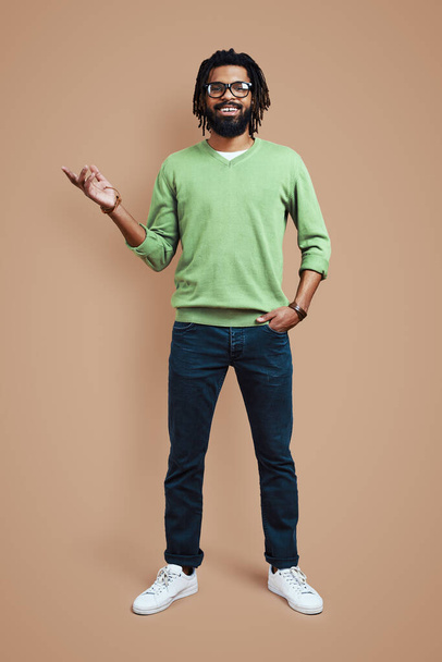 Full length of young African man in casual clothing pointing copy space and smiling while standing against brown background - Foto, Bild