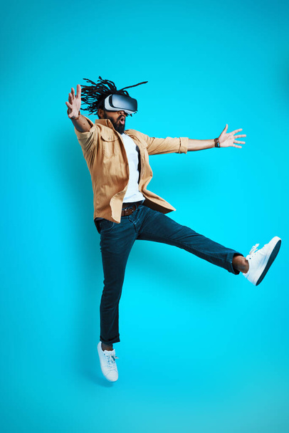 Full length of playful young African man wearing virtual reality glasses while hovering against blue background - Fotografie, Obrázek