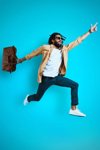 Full length of stylish young African man in casual wear smiling and carrying bag while hovering against blue background - Фото, изображение
