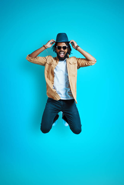 Full length of playful young African man in casual wear adjusting hat and smiling while hovering against blue background - Zdjęcie, obraz