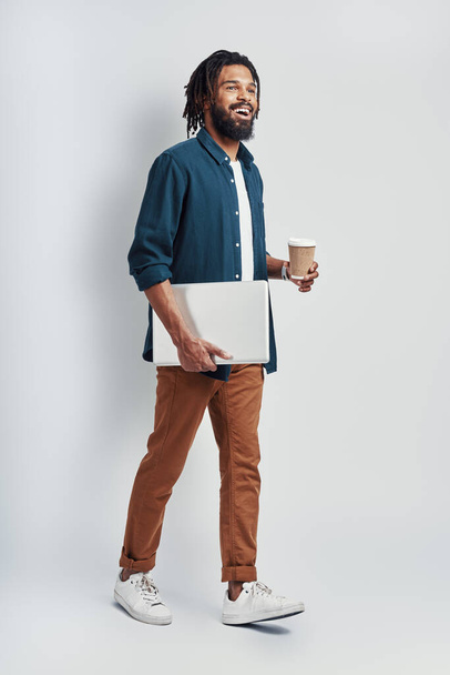 Full length of African man in eyewear holding disposable cup and carrying laptop while walking against grey background - Foto, imagen