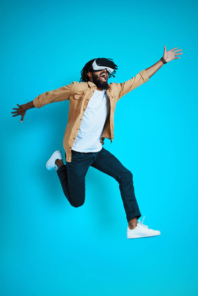 Comprimento total de brincalhão jovem africano vestindo óculos de realidade virtual enquanto paira contra fundo azul
             - Foto, Imagem