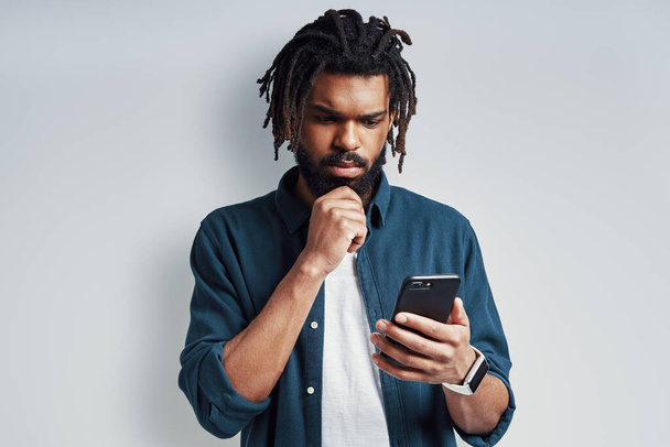 Charming young African man keeping hand on chin and using smartphone while standing against grey background - Foto, immagini