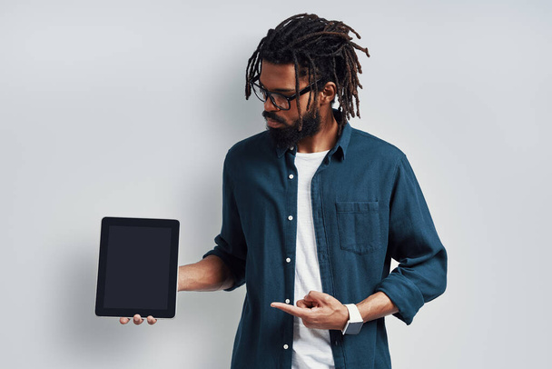 Charming young African man in eyewear pointing copy space using digital tablet while standing against grey background - Φωτογραφία, εικόνα