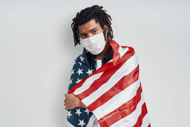 Confident young African man covered with American flag wearing medical face mask and looking at camera while standing against grey background - Φωτογραφία, εικόνα