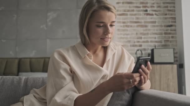 Happy attractive blonde woman using smartphone while sitting on sofa at home - Кадры, видео