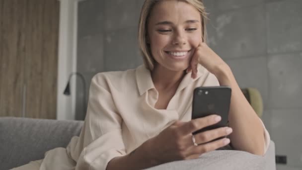Smiling attractive blonde woman using smartphone and having fun while sitting on sofa at home - Felvétel, videó