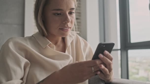 Carefree attractive blonde woman using smartphone while sitting on sofa at home - Materiał filmowy, wideo