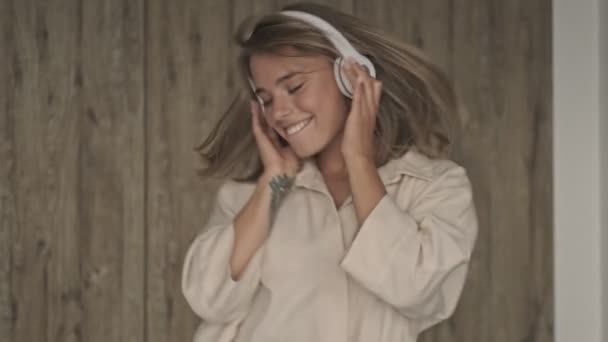 Happy attractive blonde woman in headphones listening music and dancing at home - Záběry, video