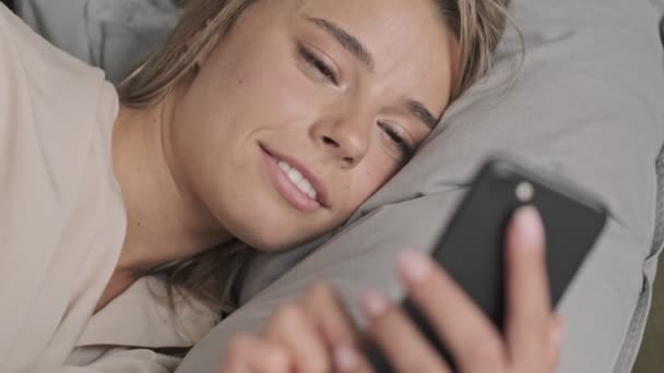 Close up view of Pleased attractive blonde woman using smartphone while lying on bed at home - Кадри, відео
