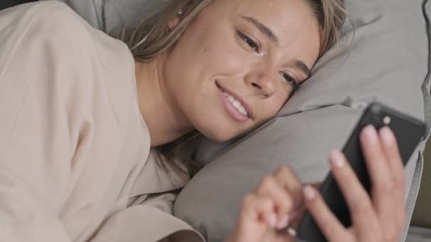 Satisfied attractive blonde woman using smartphone while lying on bed at home - Video
