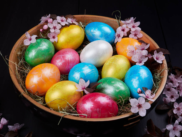 Colorful Easter eggs in a wooden bowl, on a bed of hay, with flowering branches of purple leaf plum tree       - 写真・画像