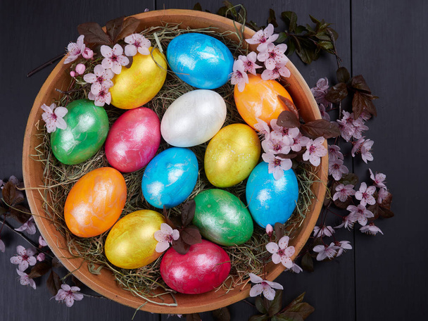 Colorful Easter eggs in a wooden bowl, on a bed of hay, with flowering branches of purple leaf plum tree       - 写真・画像