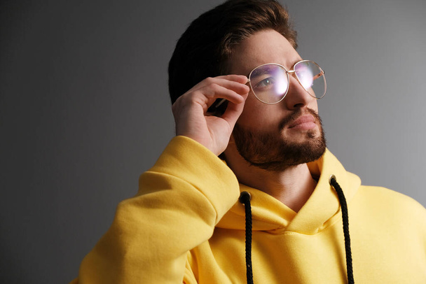 Fashionable young man 25-30 years old in sunglasses and a yellow sweatshirt posing against the background of a gray wall. - Фото, изображение