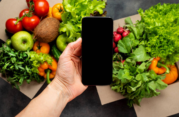 Weibliche Hand mit Smartphone und vollen Kisten mit verschiedenen frischen Bio-Gemüsen. Lieferung oder Online-Bestellung von Lebensmitteln. - Foto, Bild
