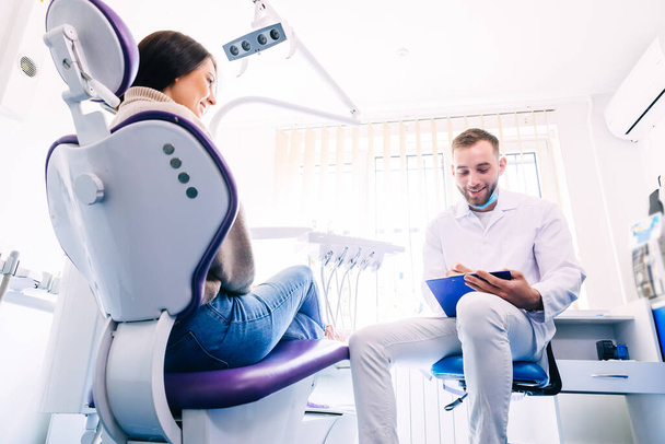 Ein lächelnder Patient sitzt auf einem Stuhl und beantwortet die Frage eines Zahnarztes. Der Zahnarzt am Arbeitsplatz führt eine Patientenbefragung durch. Geschichte nimmt Fahrt auf. - Foto, Bild