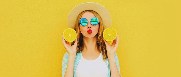 Summer portrait of young woman with fruits slice of orange blowing red lips sending sweet air kiss wearing straw hat on yellow background - Φωτογραφία, εικόνα