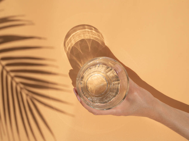 Концепция летних напитков и пляжного бара. Тень пальмового листа и женская рука держат стакан воды на бежевом фоне
 - Фото, изображение