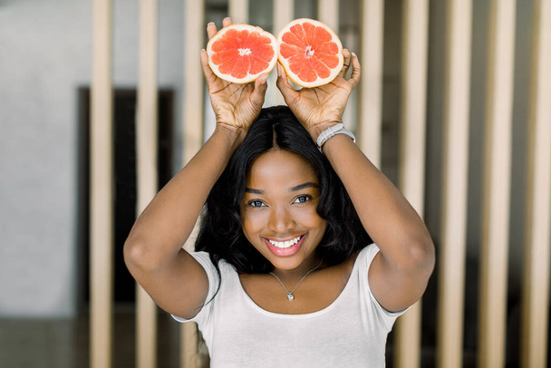Happy dark skinned African lady with dark long hair, holding halves of fresh grapefruit, above her head, smiling and having fun standing at cozy home room - Valokuva, kuva