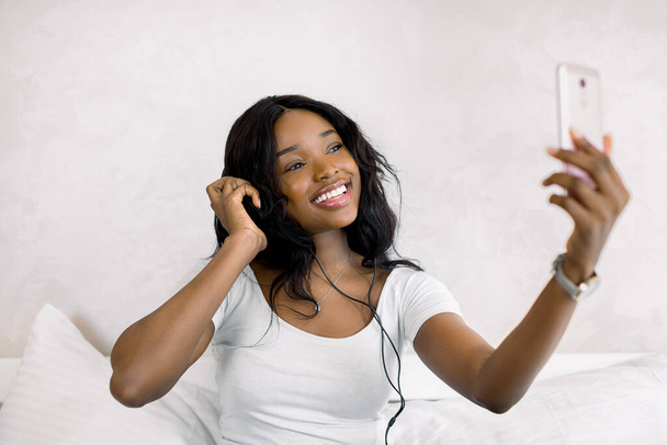 mensen, technologie en vrijetijdsbesteding. Gelukkig Afrikaans amerikaans jong vrouw zitten op bed met smartphone en koptelefoon luisteren naar muziek thuis - Foto, afbeelding