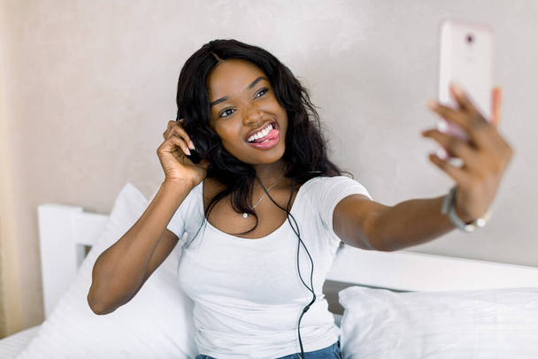 Hübsche junge Afroamerikanerin entspannt sich, hört Musik über Kopfhörer und Smartphone und macht Selfie-Fotos mit Zunge. Wohnkultur, Freizeitkonzept - Foto, Bild