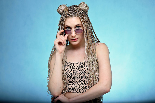 Portrait of a caucasian fashionable woman in glasses with long hair afro dreadlocks in a studio. Pretty model posing on a blue background standing right in front of the camera - Photo, Image